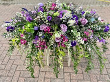 A casket spray in purple and lilac shades. Foliage drapes over the sides of the casket.