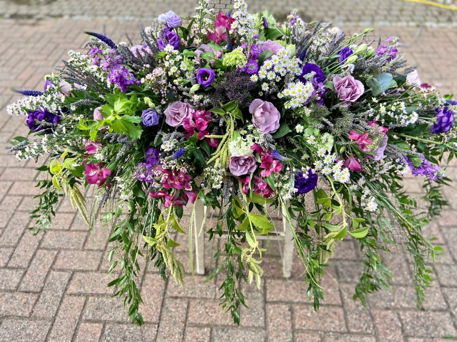 Purple and Lilac Casket Spray A casket spray in purple and lilac shades. Foliage drapes over the sides of the casket.